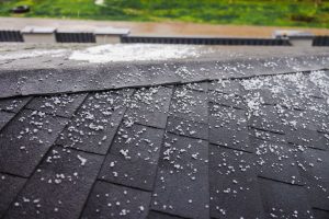 Dark shingled roof covered with scattered small hailstones during a hailstorm, with a green lawn in the background